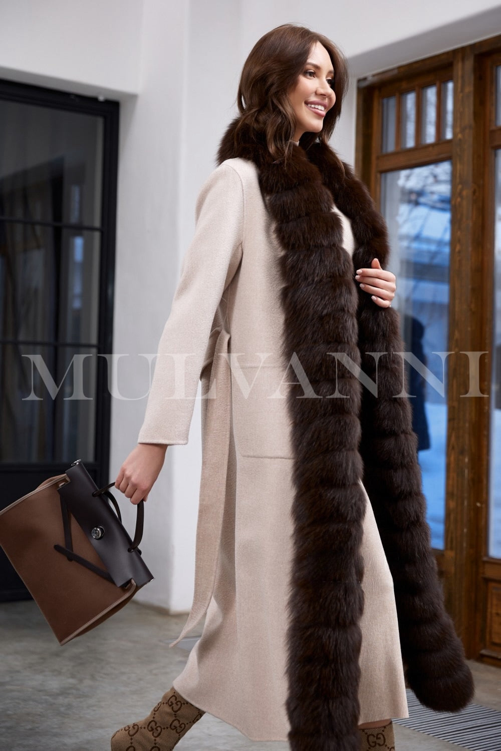 Wearing this beige double-faced cashmere coat, the woman highlights the flattering silhouette enhanced by a waist-cinching belt and plush brown fox fur detail.