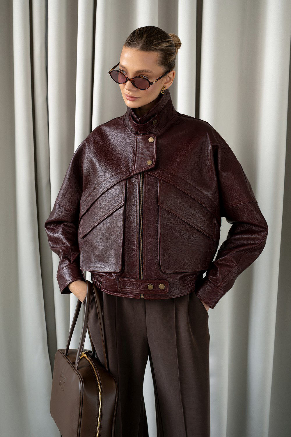 Woman in structured burgundy leather jacket styled with wide-leg trousers and sunglasses, showcasing bold luxury outerwear.