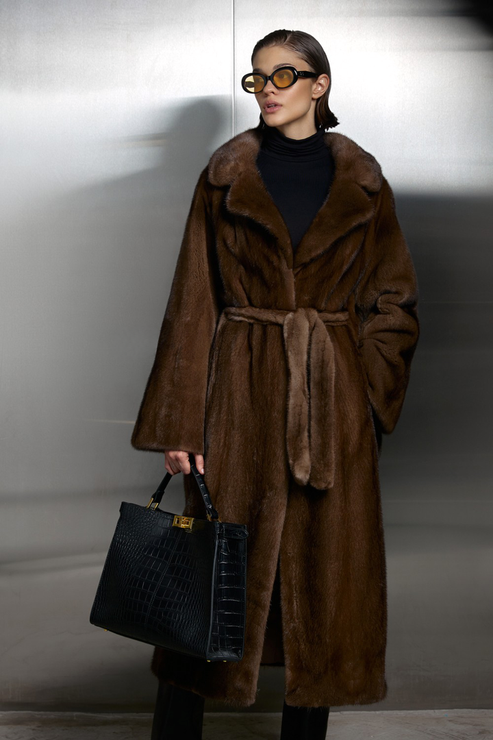 Luxurious long genuine brown mink fur coat with a waisted belt, worn with a stylish black bag.