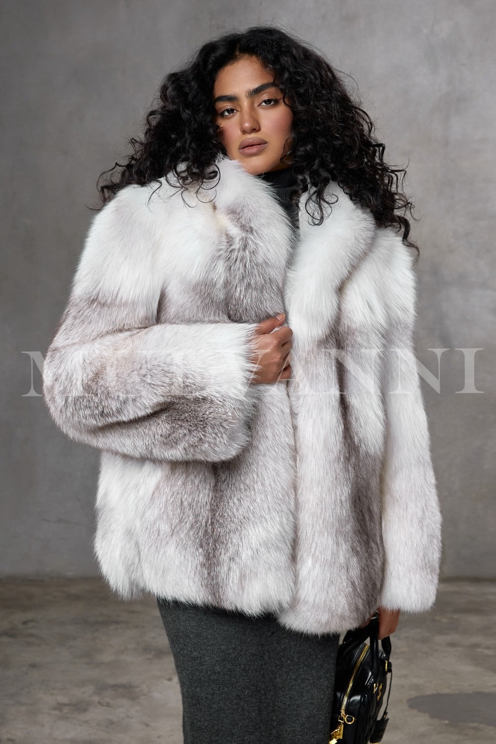 A woman wears a meticulously handcrafted full-pelt Arctic fox fur coat, featuring exceptional softness, volume, and a loose fit with a plush hood that envelops her in luxurious warmth and elegant style.