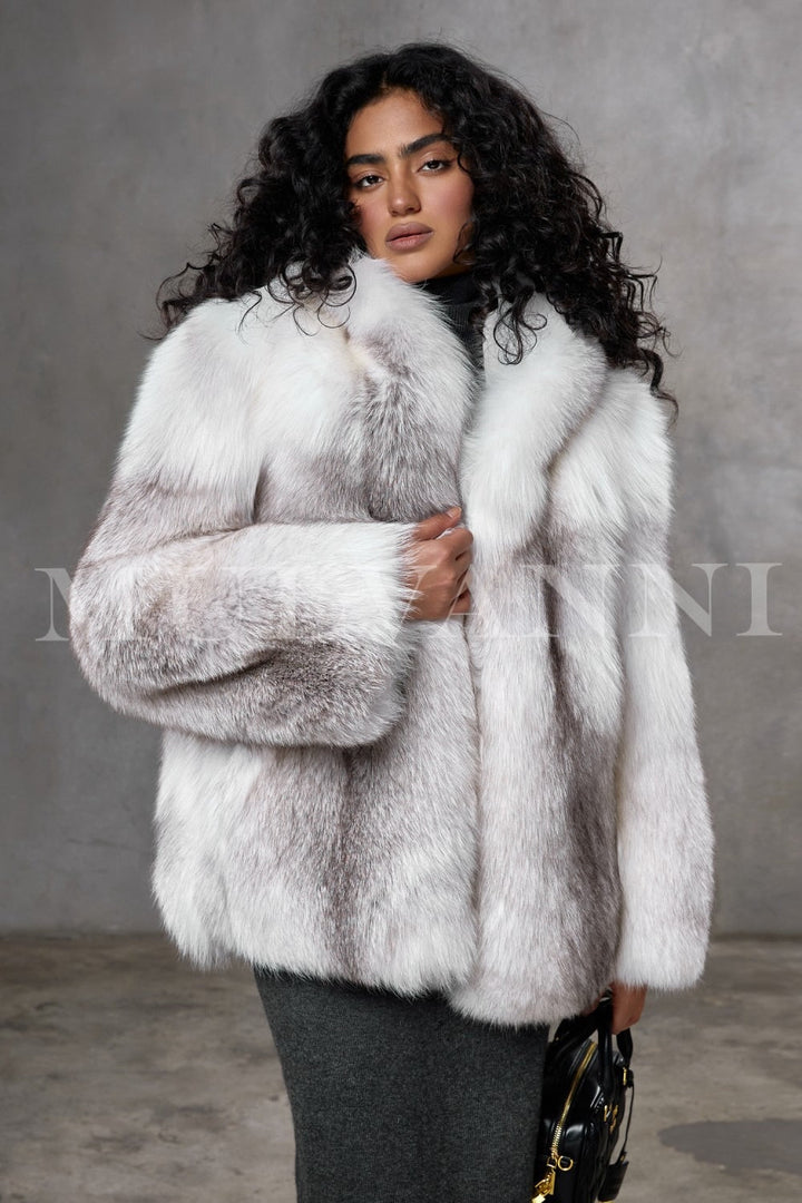 A woman wears a meticulously handcrafted full-pelt Arctic fox fur coat, featuring exceptional softness, volume, and a loose fit with a plush hood that envelops her in luxurious warmth and elegant style.