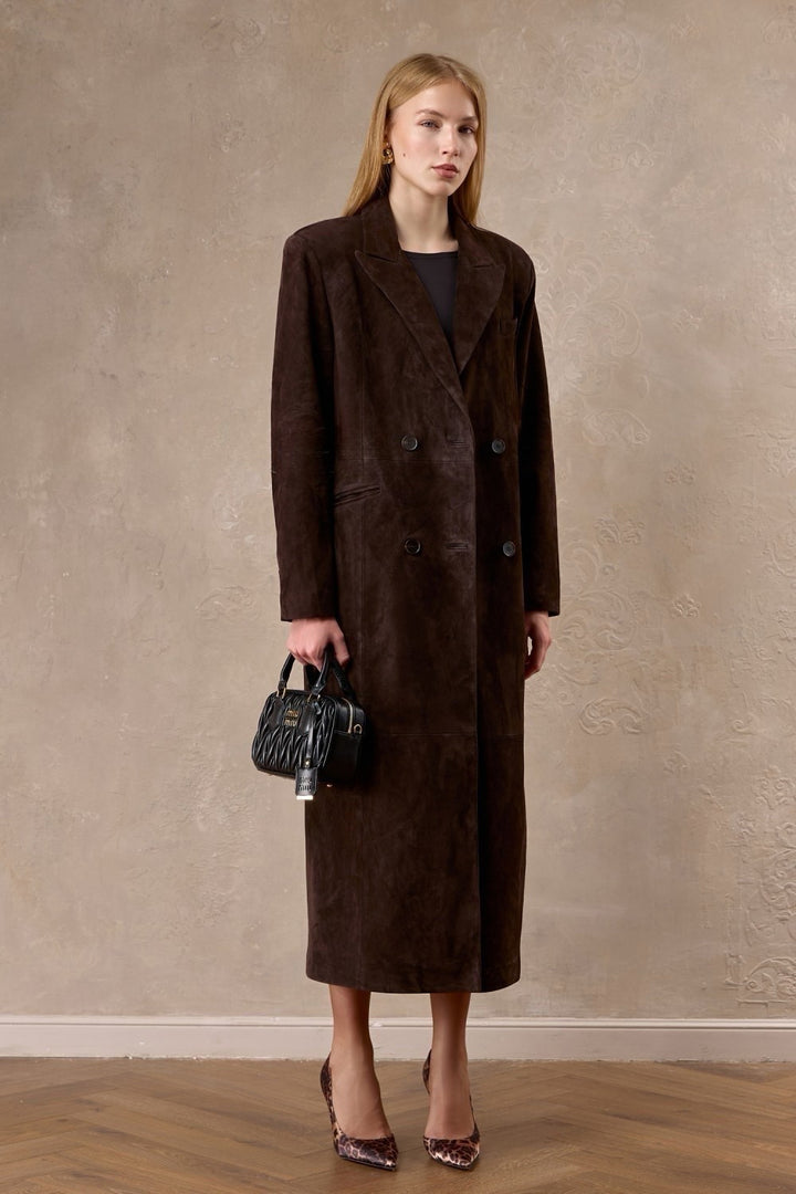 A woman is seen wearing a long chocolate suede coat with sculpted shoulders and a belted waist, exuding quiet confidence and elegance.