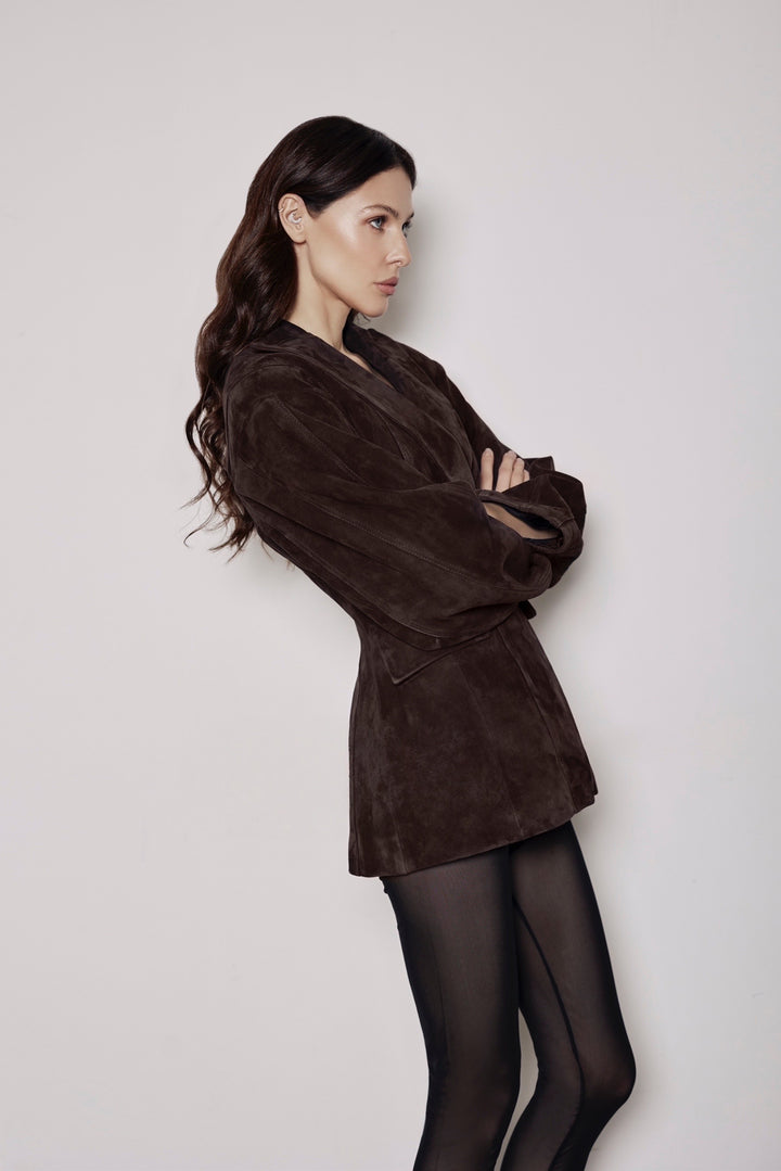 A woman wearing a brown suede blazer with a cinched waist and black tights, standing confidently with a sleek silhouette that blends softness and structure.