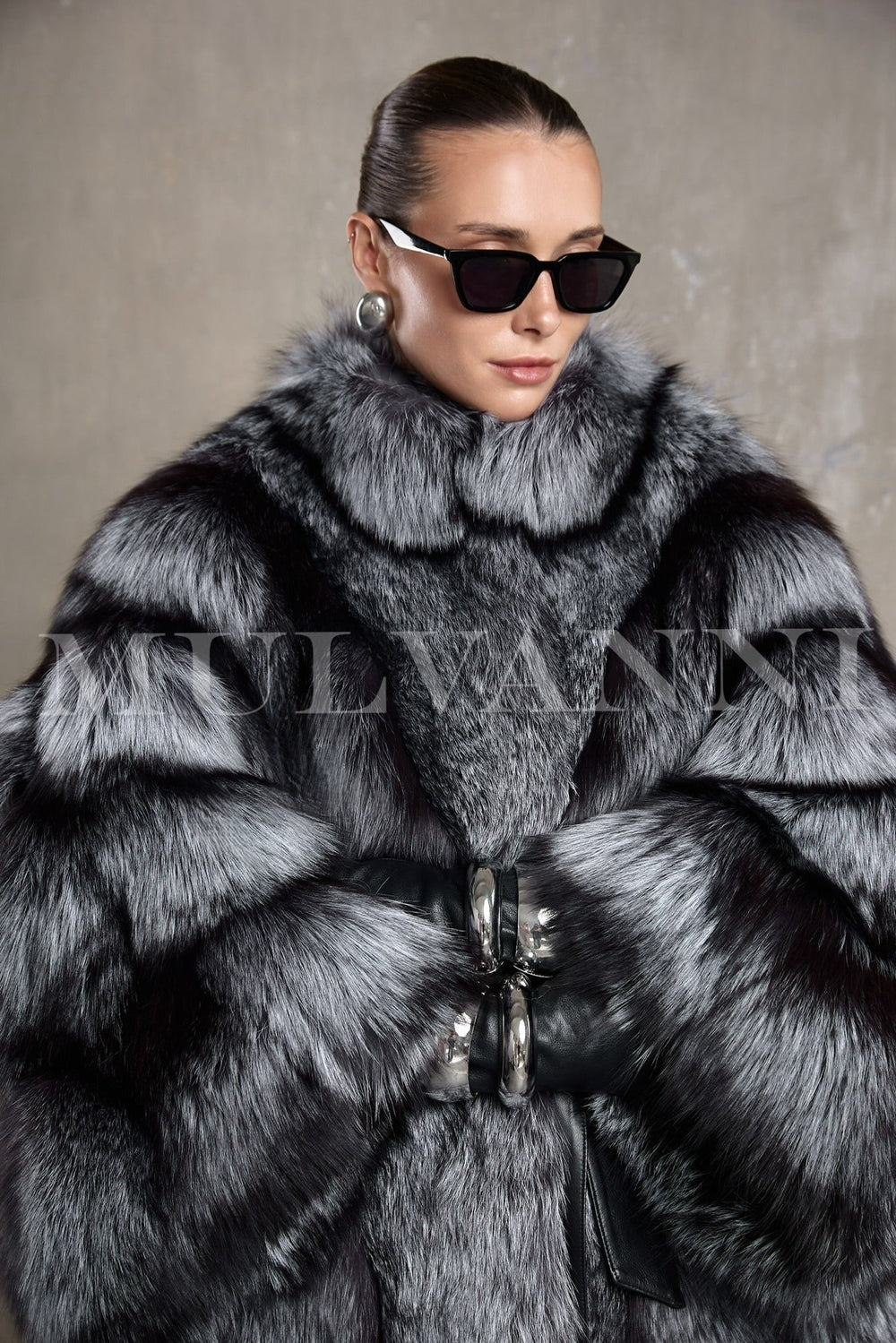 A woman wears an elegant full-length silver fox fur coat with a voluminous silhouette and leather belt that cinches the waist.