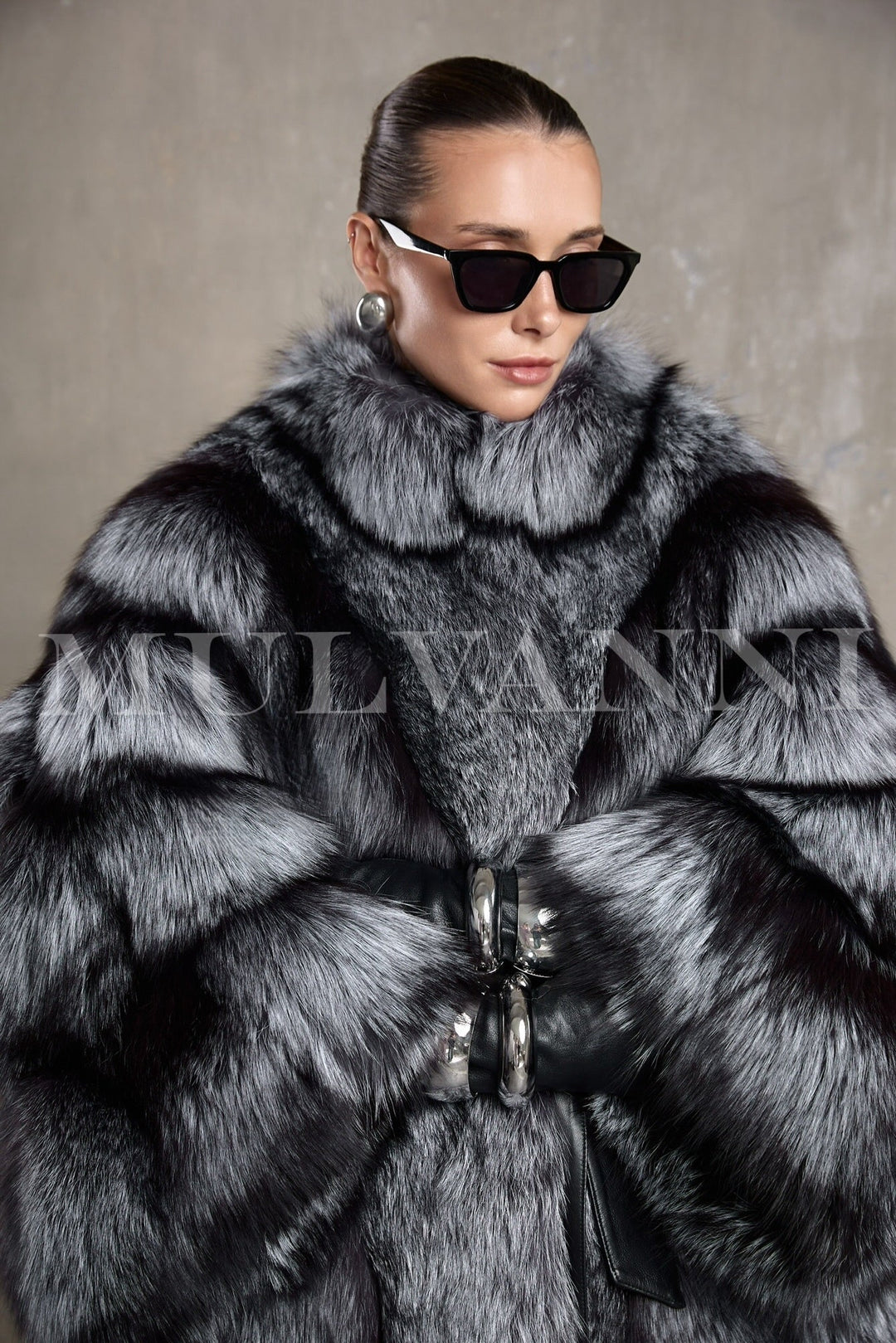 A woman wears an elegant full-length silver fox fur coat with a voluminous silhouette and leather belt that cinches the waist.