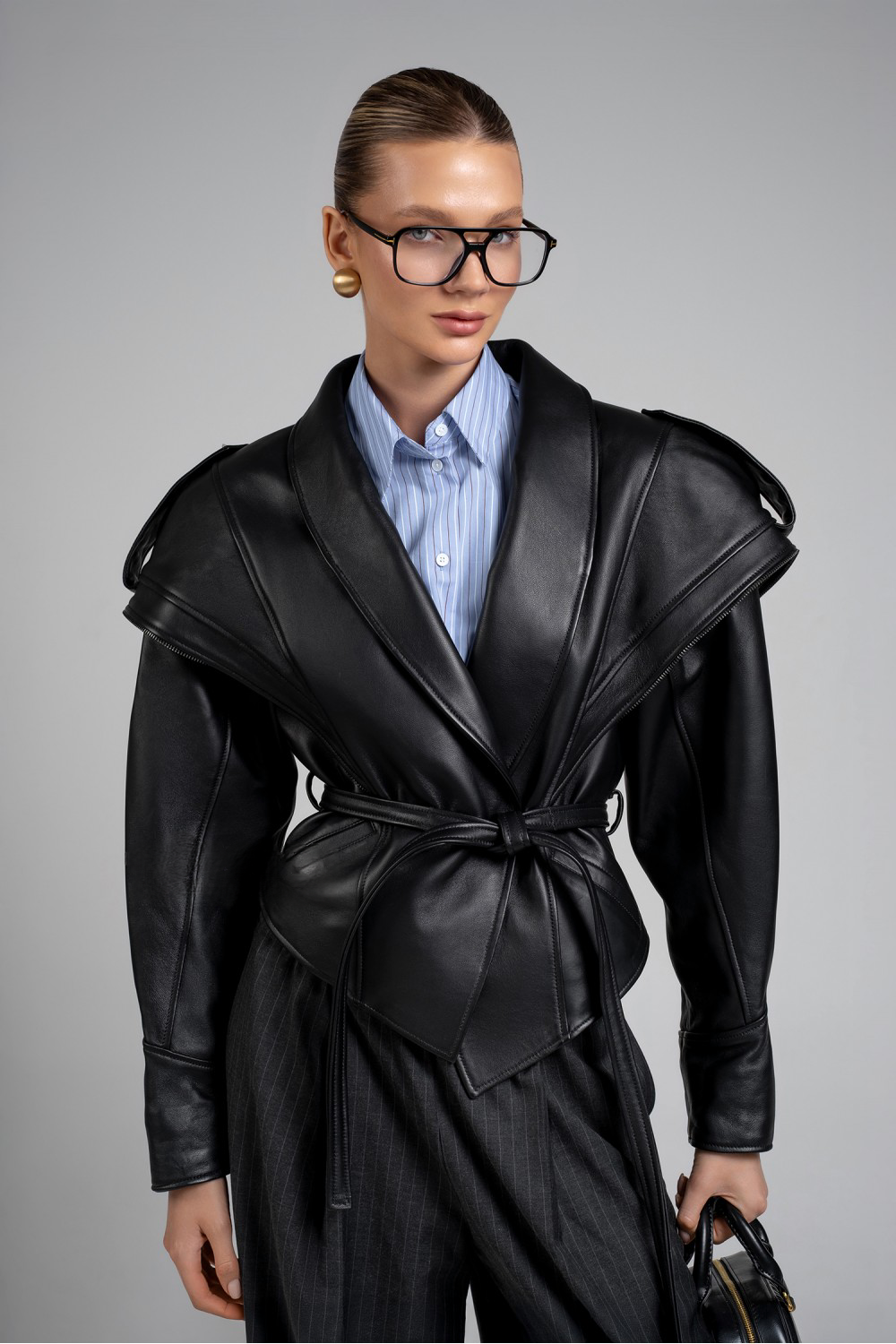 A woman wearing a Jacket, made from supple black leather with bold architectural shoulders, an asymmetric front zip, and a cinched waist tied with a sleek leather belt for a powerful, modern look.