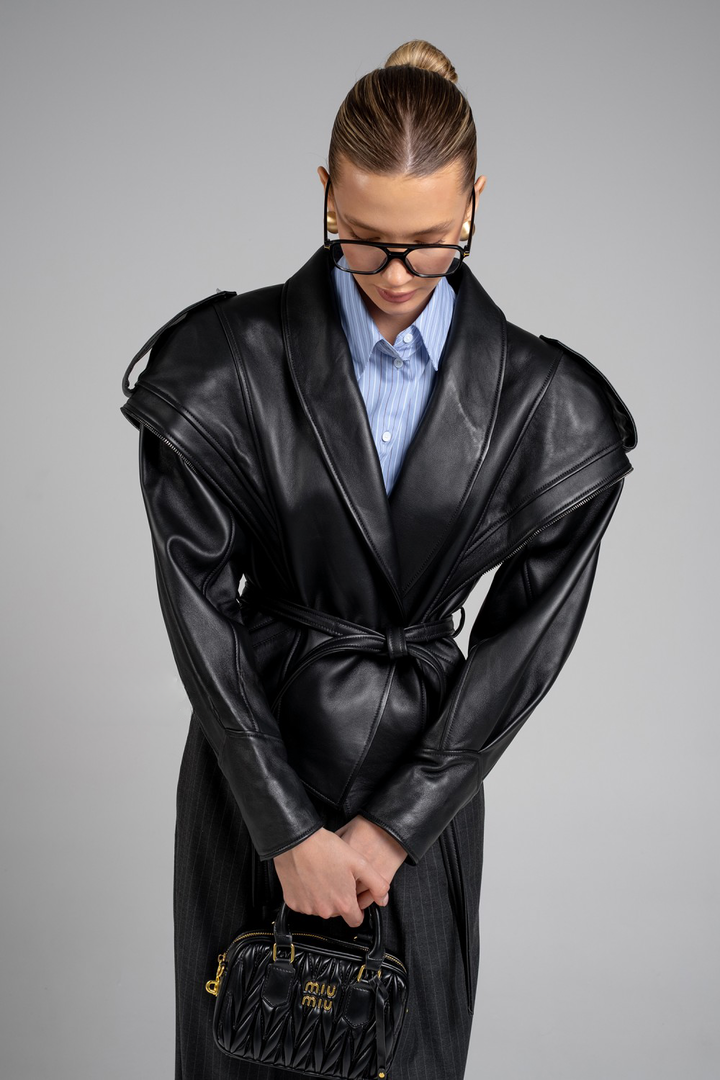A woman models a striking black leather jacket featuring architectural shoulders, a sharp asymmetric zip, and a belted waist that accentuates the figure with an edgy, feminine twist.