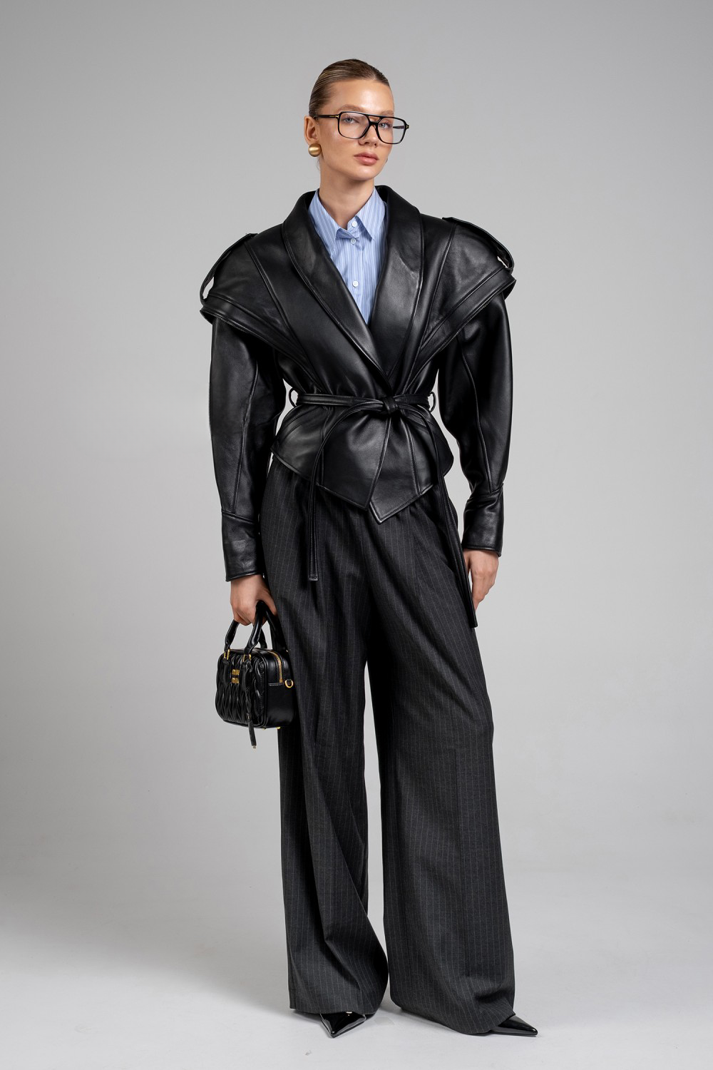 A woman wearing a bold black leather jacket with sculpted shoulders, an asymmetric front zip, and a cinched waist tied with a sleek leather belt, creating a modern, empowered silhouette.