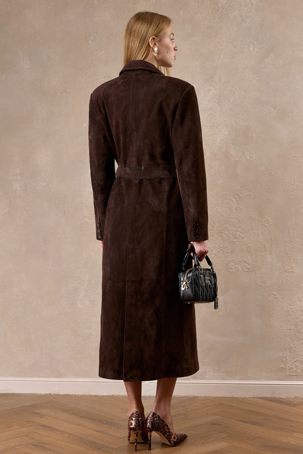 Draped in a chocolate suede coat, a woman showcases a tailored fit with wide shoulders and a cinched waist, blending sophistication with strength.