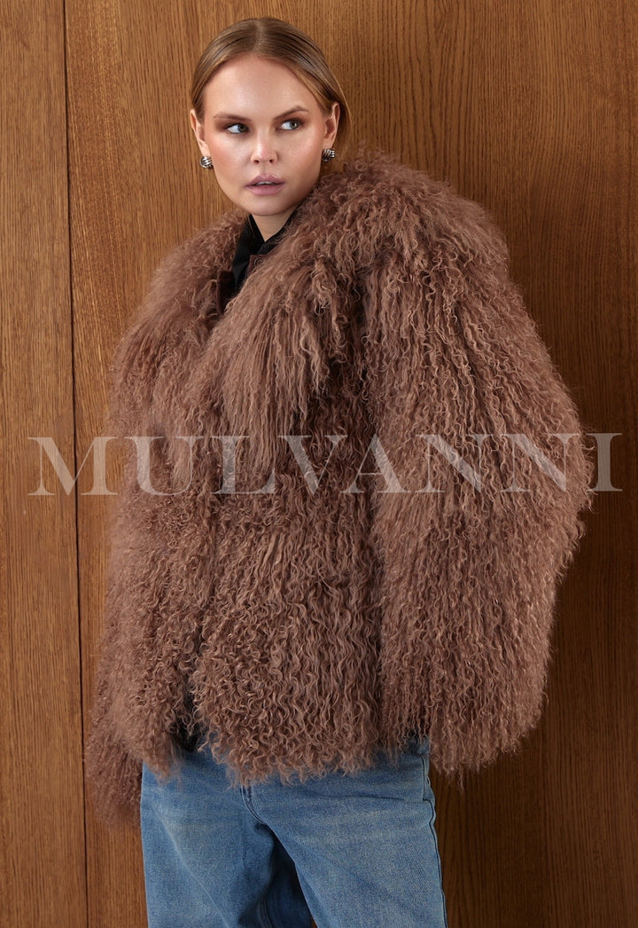 The woman models a one-piece brown Mongolian coat, featuring soft, curly fur, side slit pockets, and a flowing, opulent look.