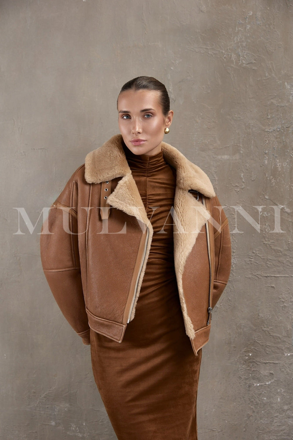 Model wearing a brown sheepskin leather bomber with silver zippers and double-breasted front, layered over a winter sweater.