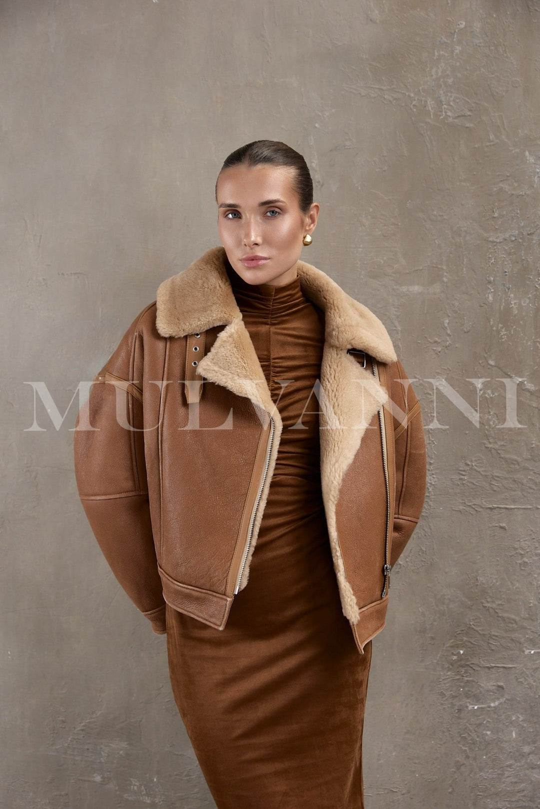 Model wearing a brown sheepskin leather bomber with silver zippers and double-breasted front, layered over a winter sweater.
