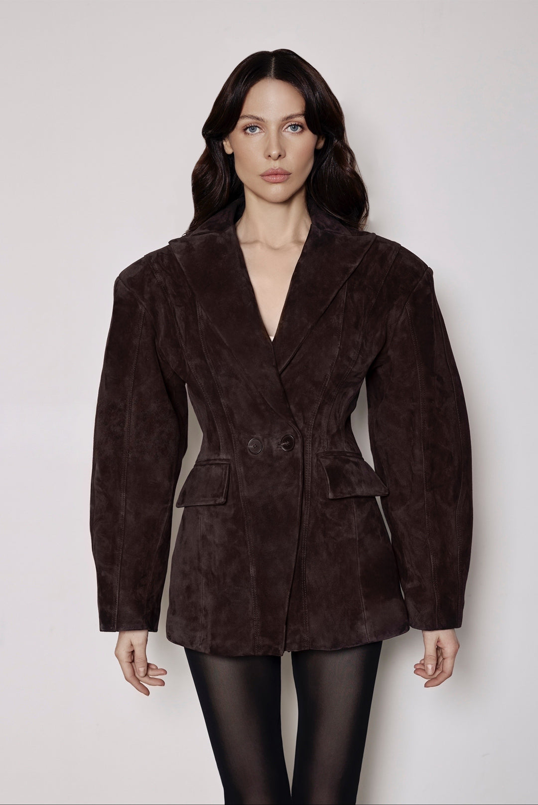 A woman wearing a brown suede blazer tailored with a cinched waist, paired with black tights, blending soft suede with structured elegance for a polished feminine look.