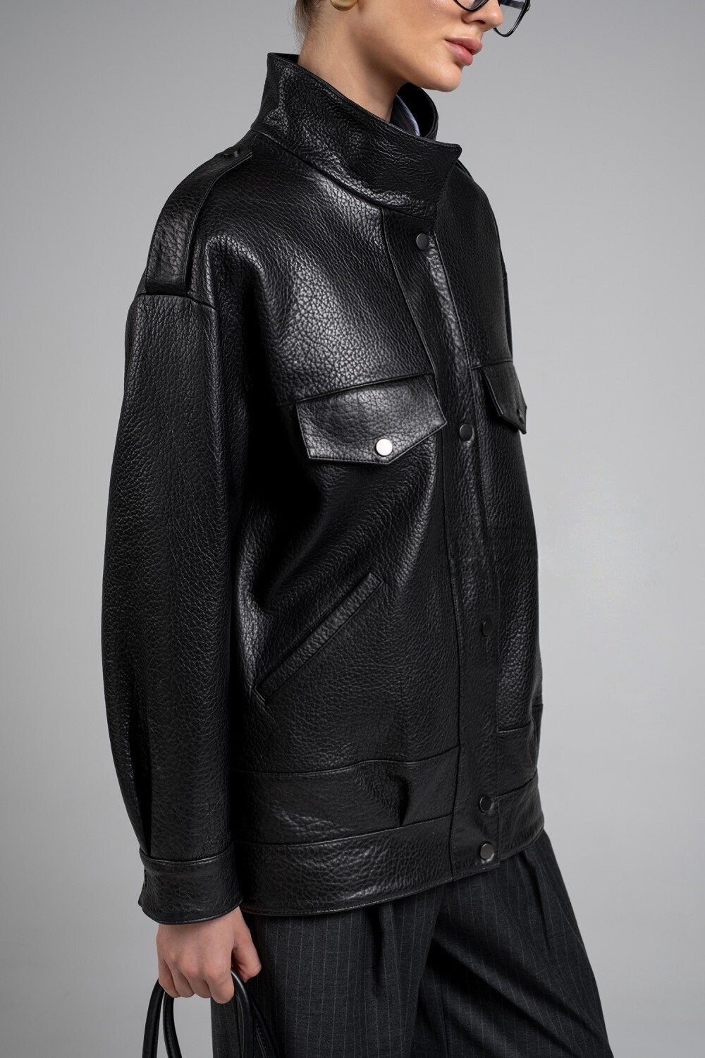 A woman wearing a modern black genuine leather jacket with a sleek high collar and tailored fit, paired with fashionable glasses and a black leather bag for effortless winter style.