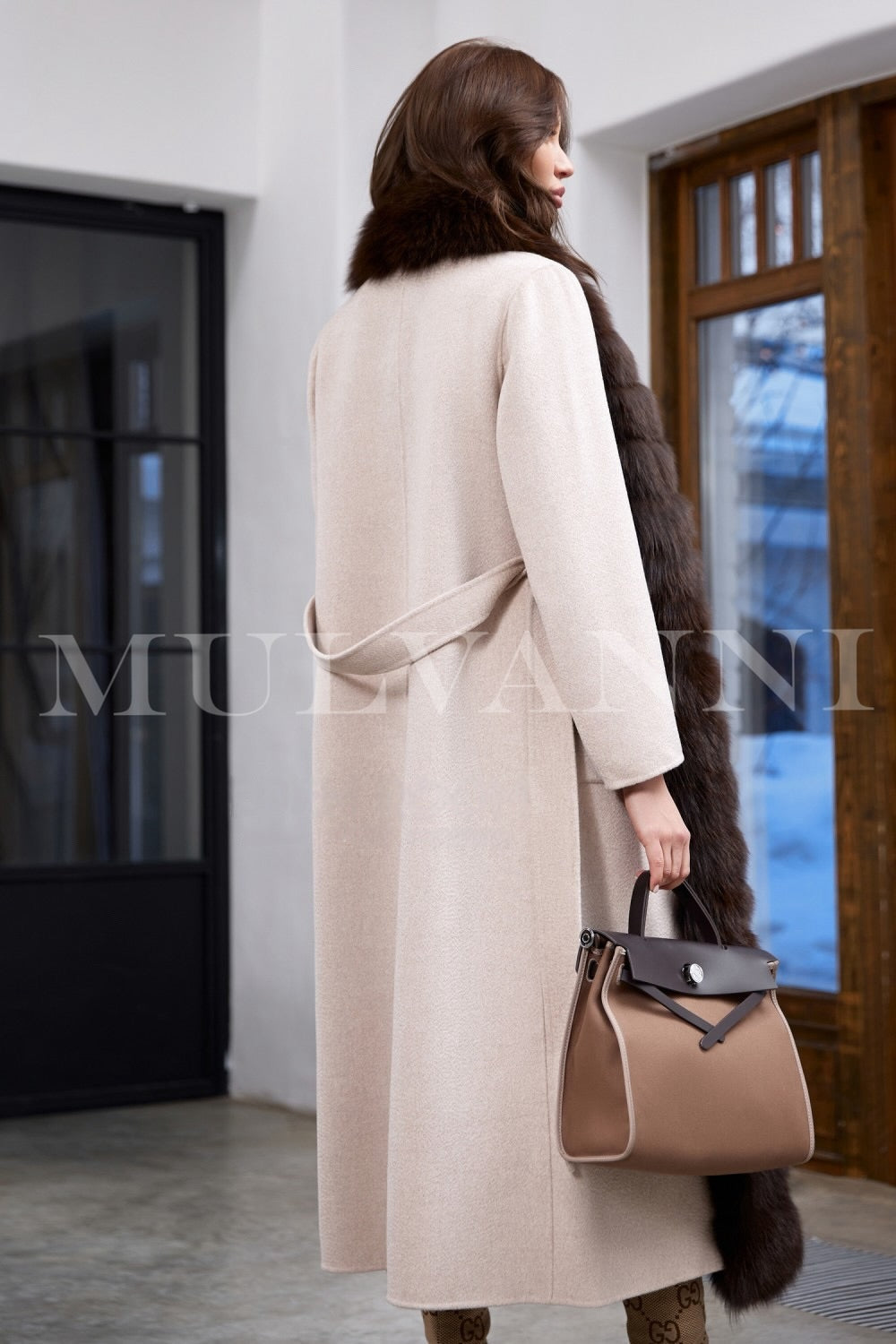 A woman elegantly dons a beige cashmere coat, featuring an impeccable fit, a cashmere belt at the waist, and rich brown fox fur trim for added warmth and style.