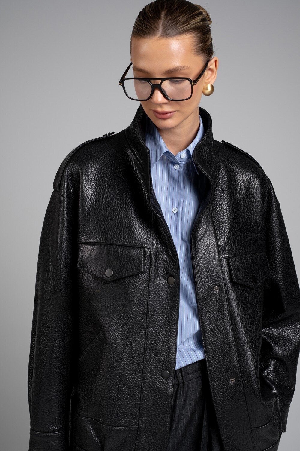 A woman models a black leather jacket with clean lines and a high neck, accessorized with elegant glasses and a black leather bag, creating a versatile and stylish look.