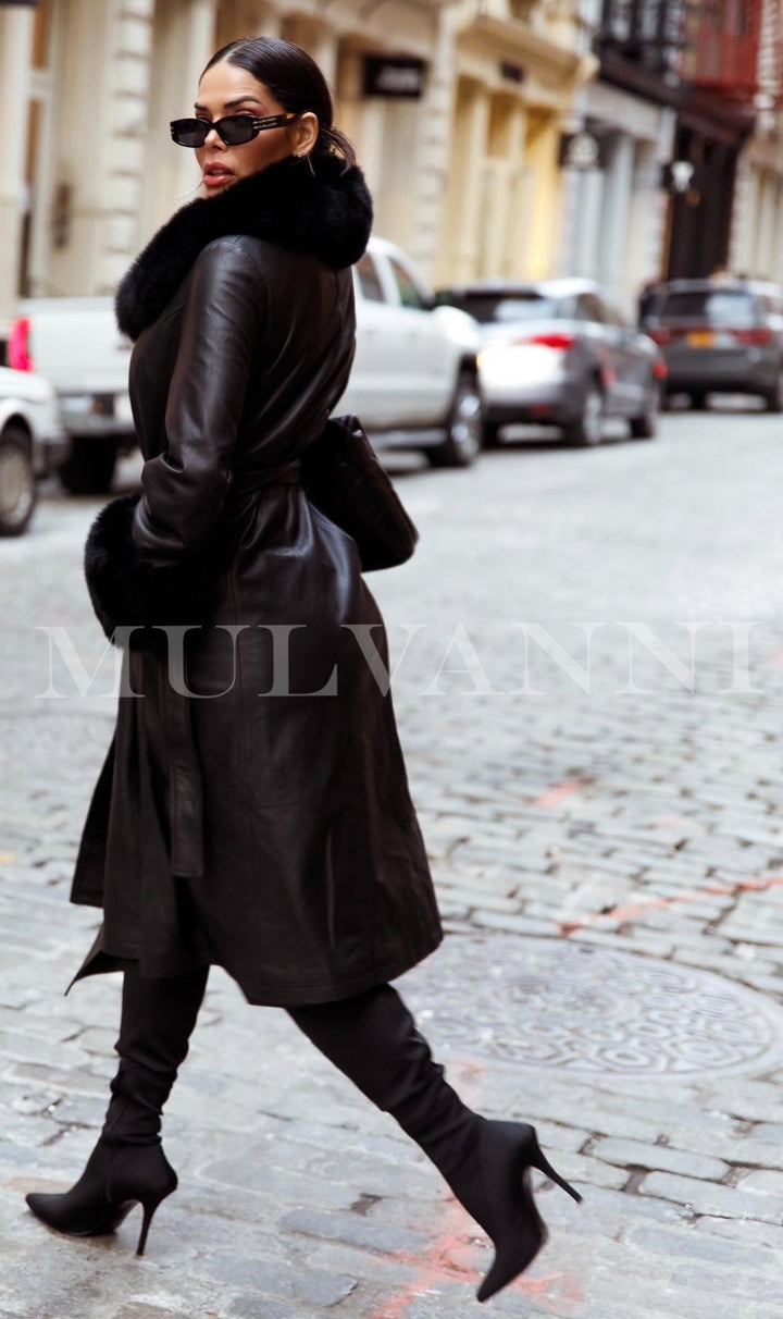 A fashionable woman models a black leather trench coat with large black fox fur accents on the collar and cuffs, blending luxury and warmth for a timeless and striking outfit.