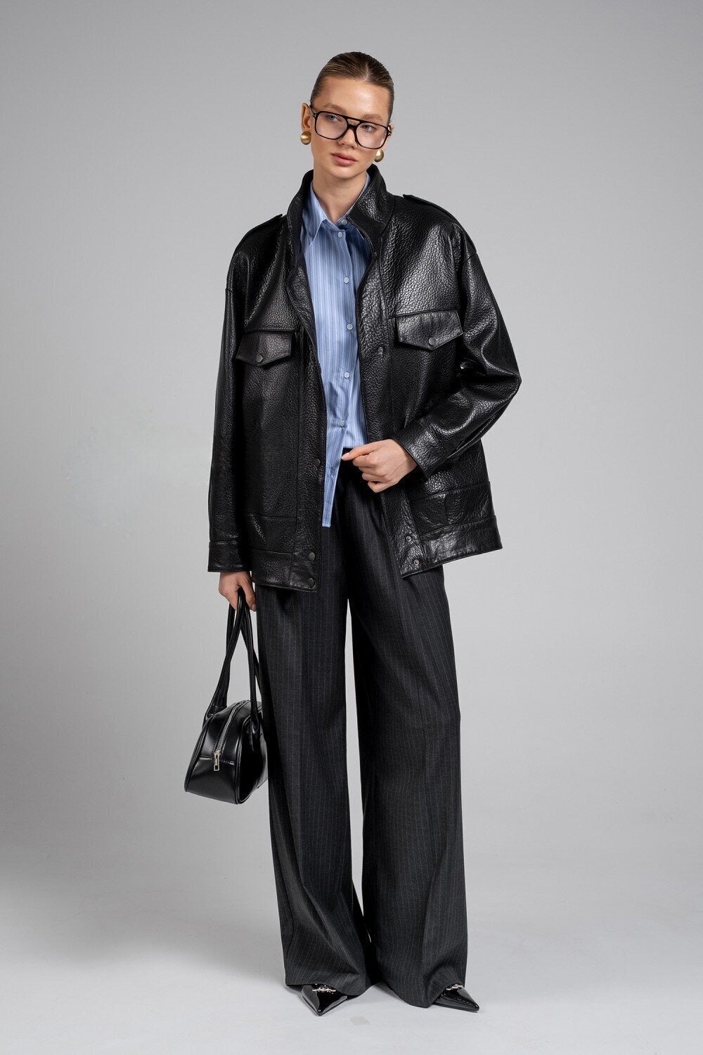 A woman models a tailored black leather jacket featuring a minimalist silhouette and high collar, wearing glasses and holding a black leather bag for a polished day-to-night outfit.