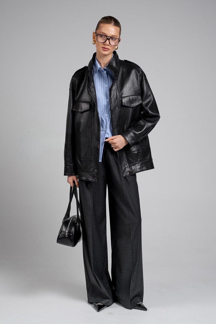 A woman models a tailored black leather jacket featuring a minimalist silhouette and high collar, wearing glasses and holding a black leather bag for a polished day-to-night outfit.