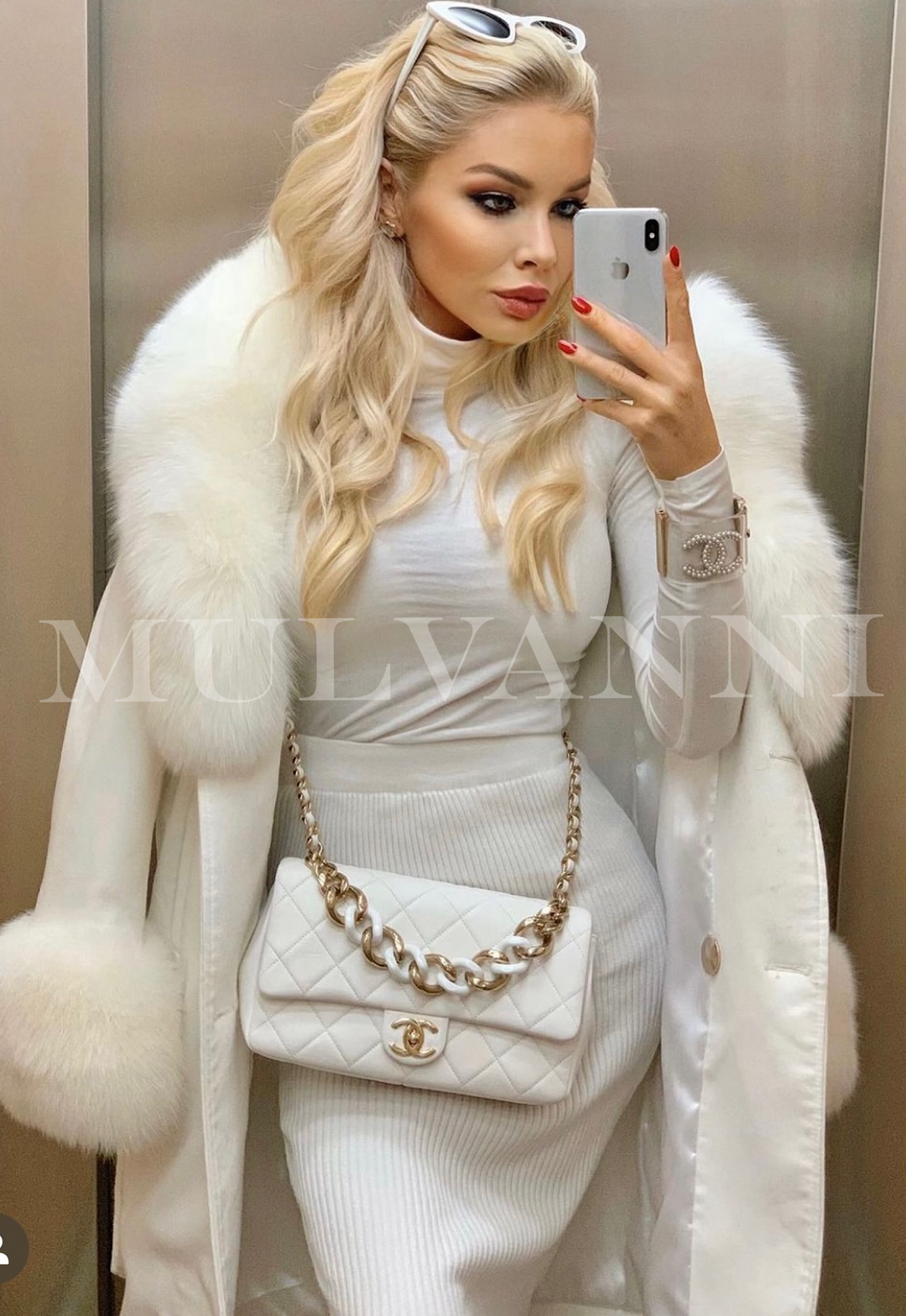 A woman wearing a glamorous off white cashmere coat with large dramatic white fox fur collar and fur cuffs combined with a white bag. 