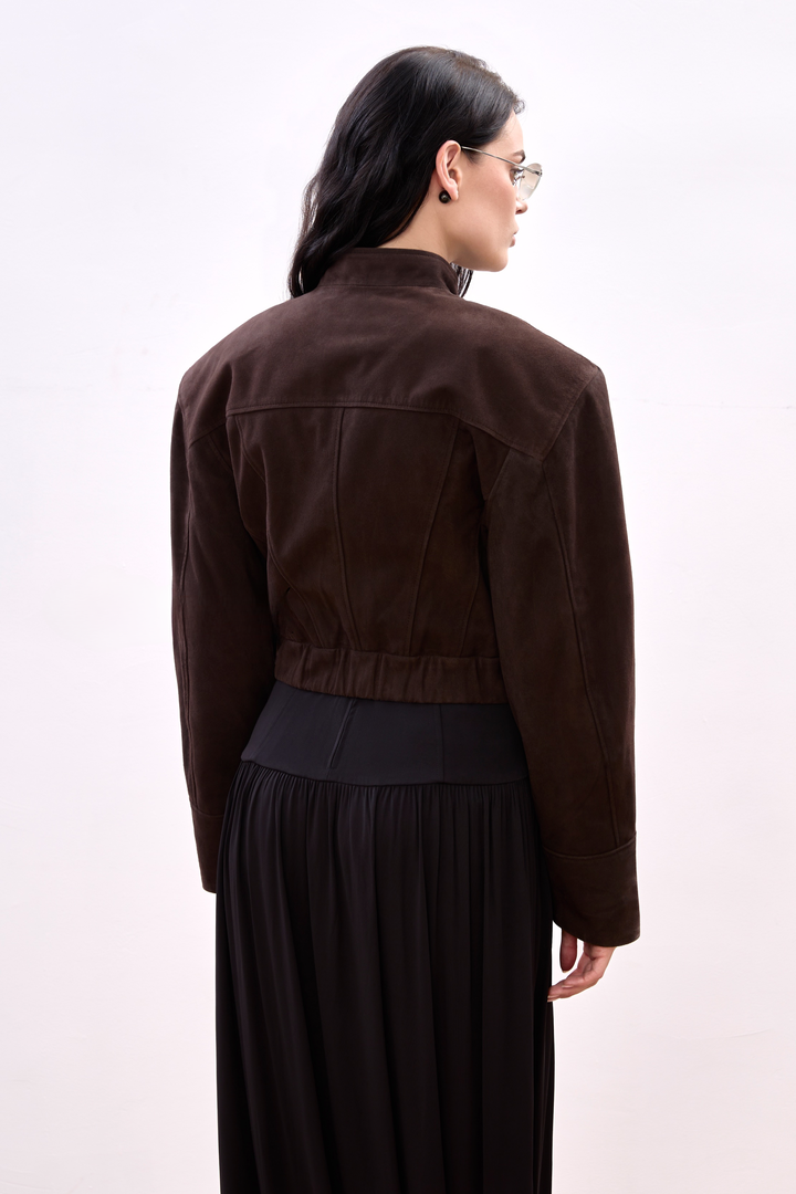 A woman models a deep espresso suede cropped jacket with a high collar and hidden front closure, blending modern minimalism with a powerful, structured silhouette.
