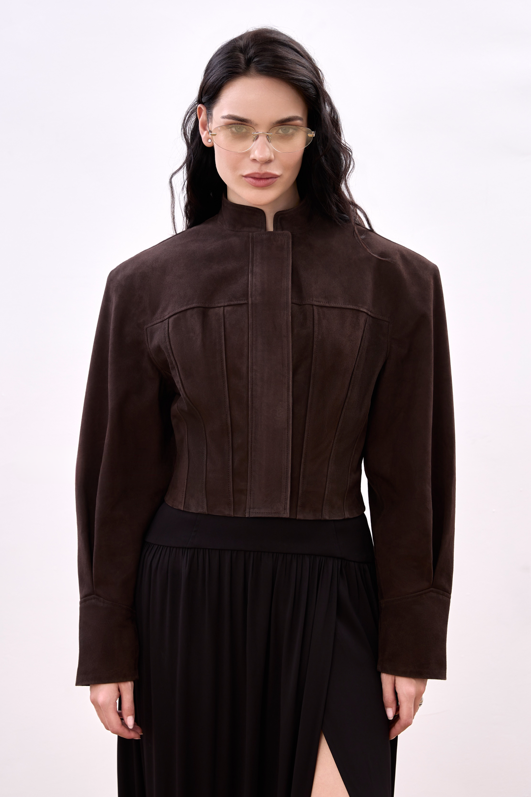 A woman wearing a sharp, espresso-colored suede jacket with a cropped silhouette, tailored to accentuate the waist and shoulders with clean, minimalist lines.