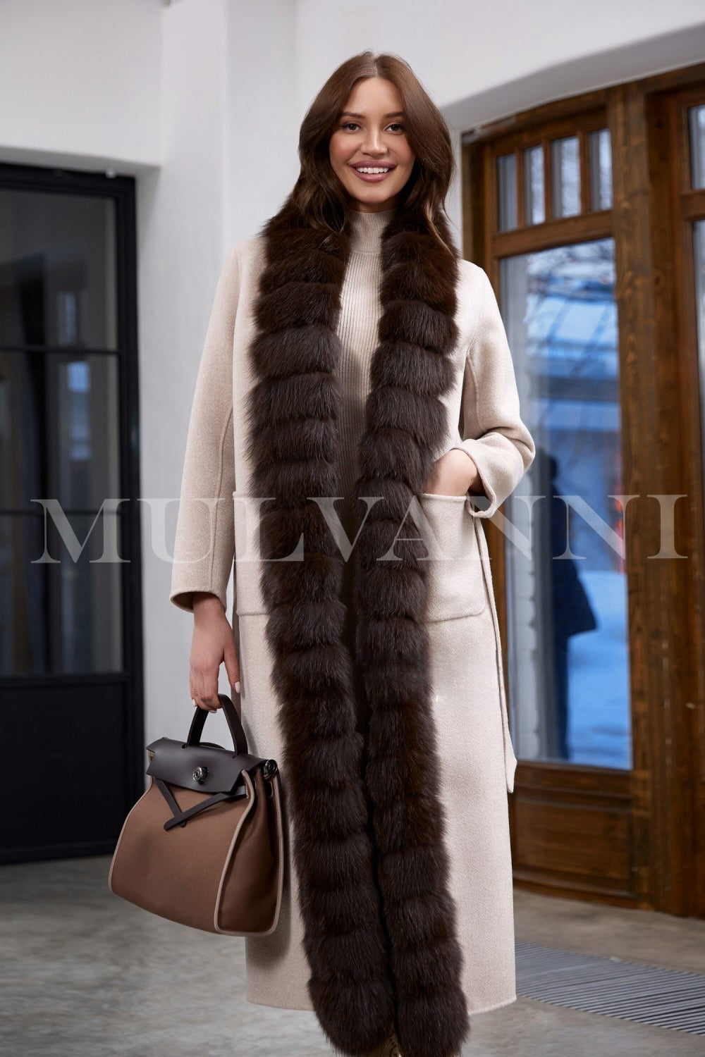 The woman wears a structured beige cashmere coat with a refined fit, complemented by a matching belt and soft brown fox fur trim adding timeless sophistication.