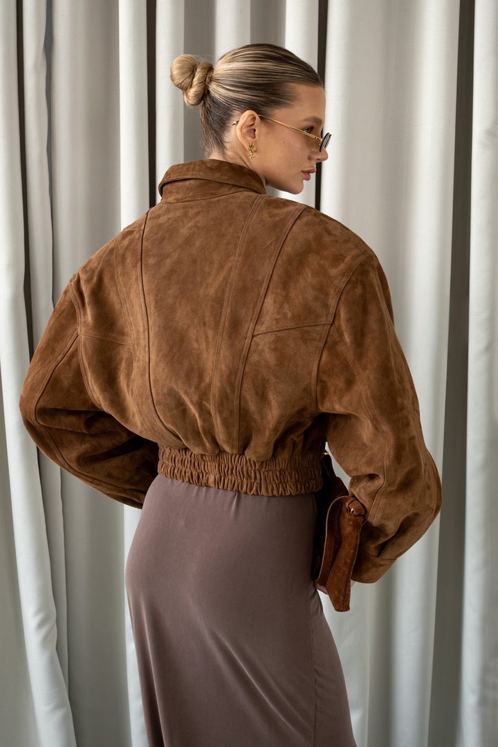 A woman models a luxurious cropped bomber jacket in genuine whiskey-hued suede, blending classic warmth with contemporary sophistication and versatile seasonal style.