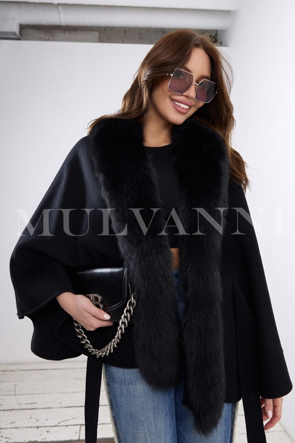 A woman wears a classic black cashmere cape trimmed with genuine fox fur, effortlessly layering it over her outfit for a chic and sophisticated look suitable year-round.