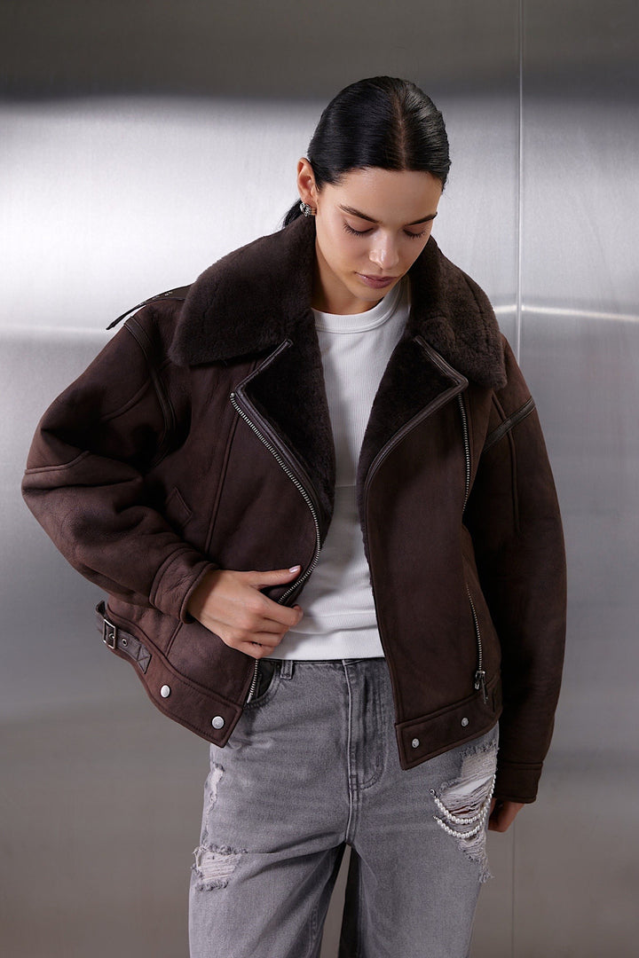 A woman wearing a classic brown aviator leather bomber jacket crafted from heavyweight sheepskin, featuring cozy shearling lining and a cropped, feminine twist on a sporty oversized style.