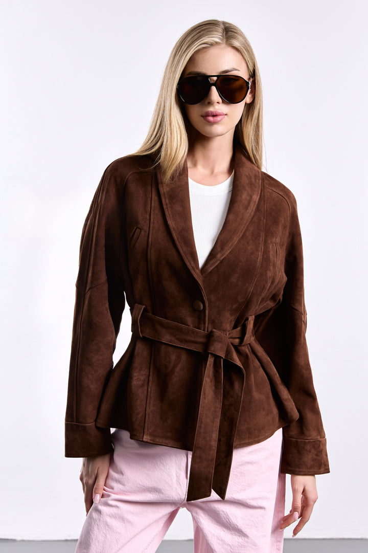 A woman wearing a brown suede jacket with a cinched belted waist, featuring a relaxed yet tailored silhouette that blends modern structure with timeless elegance.