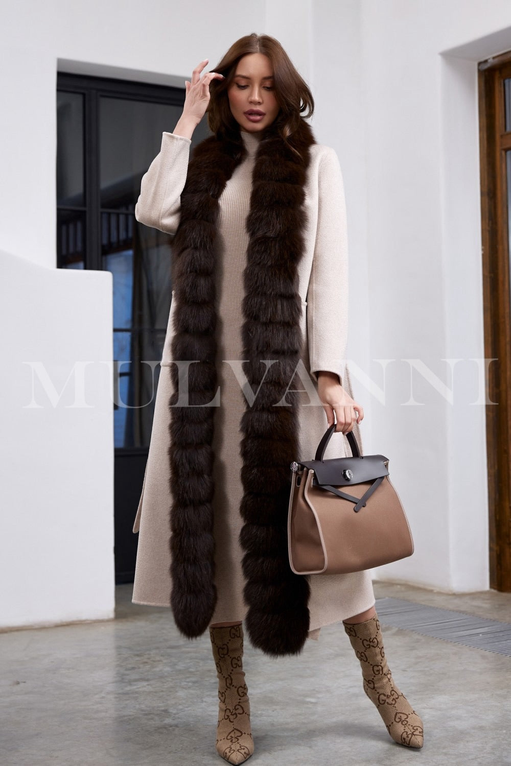 Elegantly dressed, the woman showcases a beige cashmere coat with clean lines, a cinched waist belt, and luxurious brown fox fur cascading along the front.