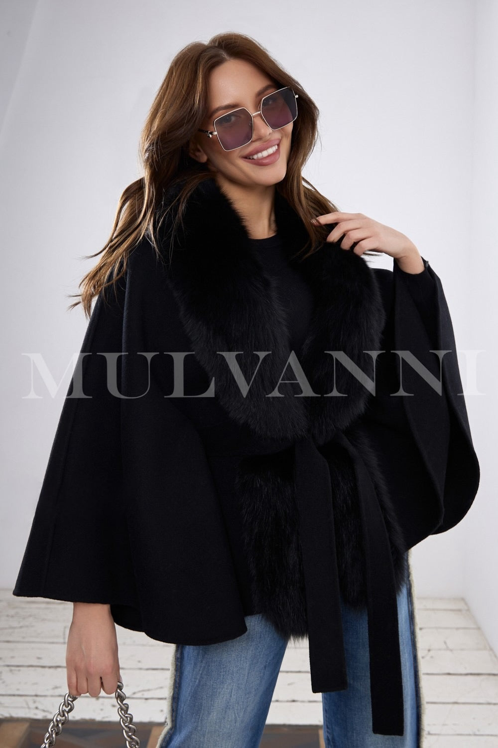 Draped in a stylish black cashmere cape with soft fox fur trim, a woman showcases timeless elegance, perfect for layering over sweaters and elevating any ensemble.