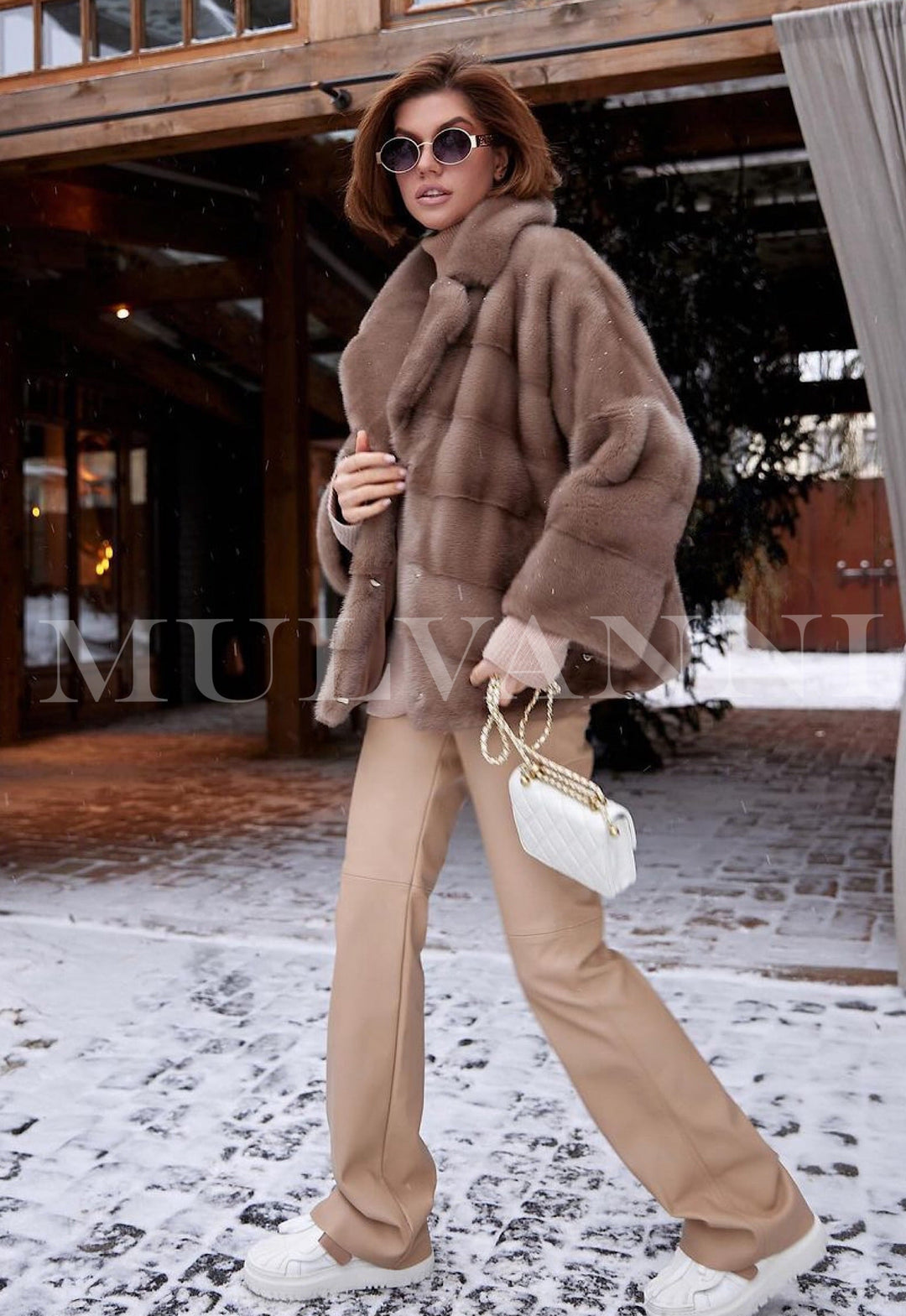 A woman wearing a stylish short brown mink jacket, paired with bold sunglasses. The soft mink fur and modern cut provide a polished and fashionable ensemble.