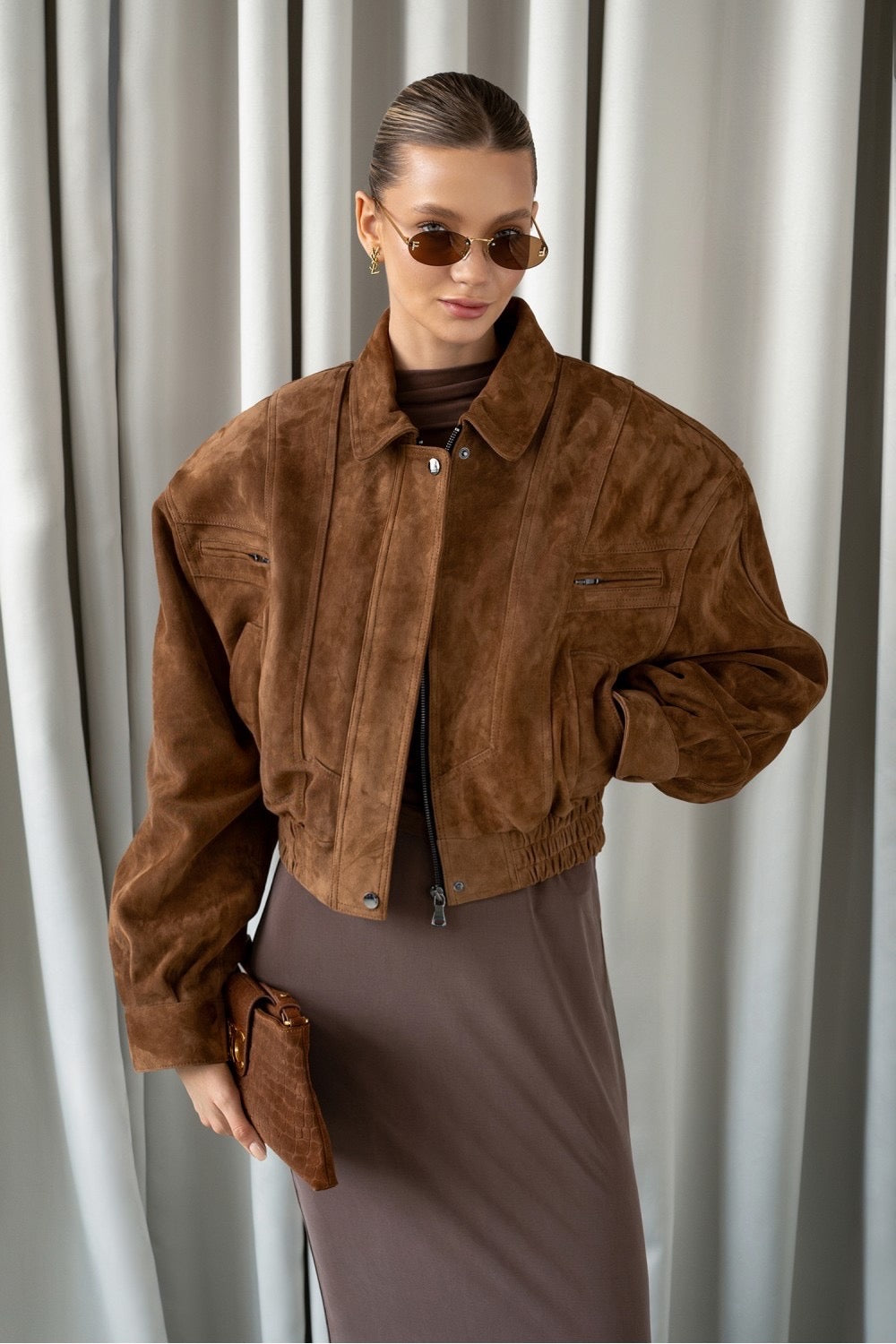A woman wearing a cropped bomber jacket crafted from ultra-soft genuine suede in a rich whiskey hue, featuring a modern waist-length silhouette that blends timeless appeal with contemporary edge.