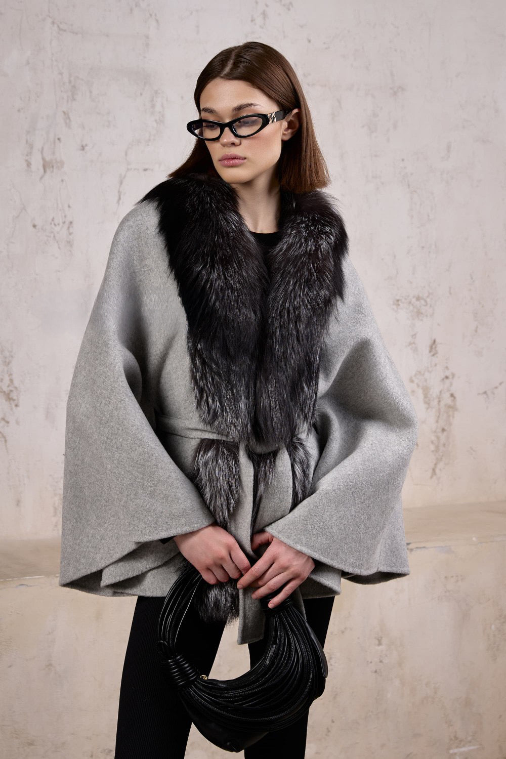 A woman wears a classic cashmere cape trimmed with genuine silver fox fur, effortlessly layering it over her outfit for stylish year-round elegance.