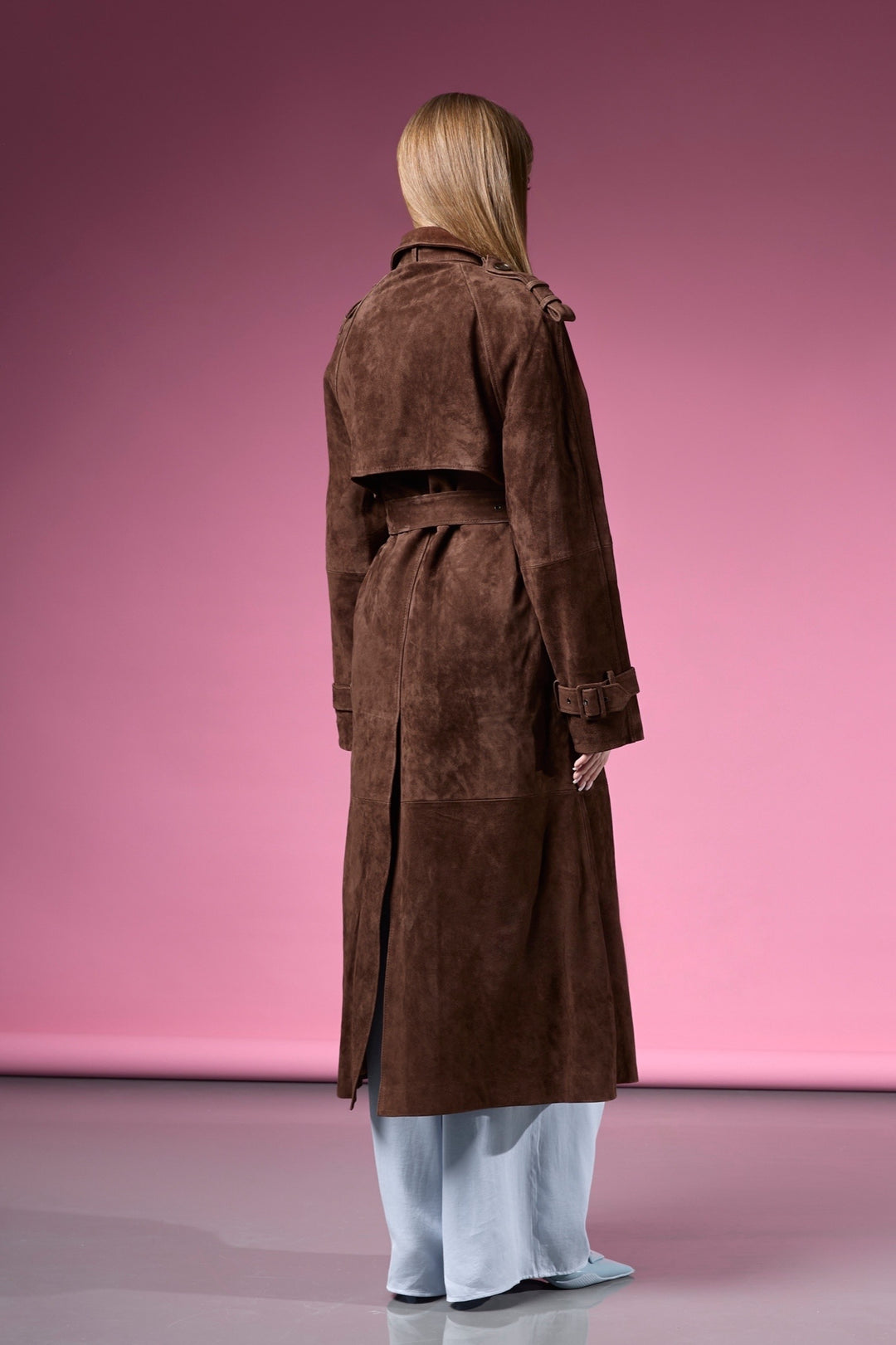 A woman models a chocolate brown trench coat in premium suede leather, designed with a figure-flattering shape and a sleek double-breasted closure.
