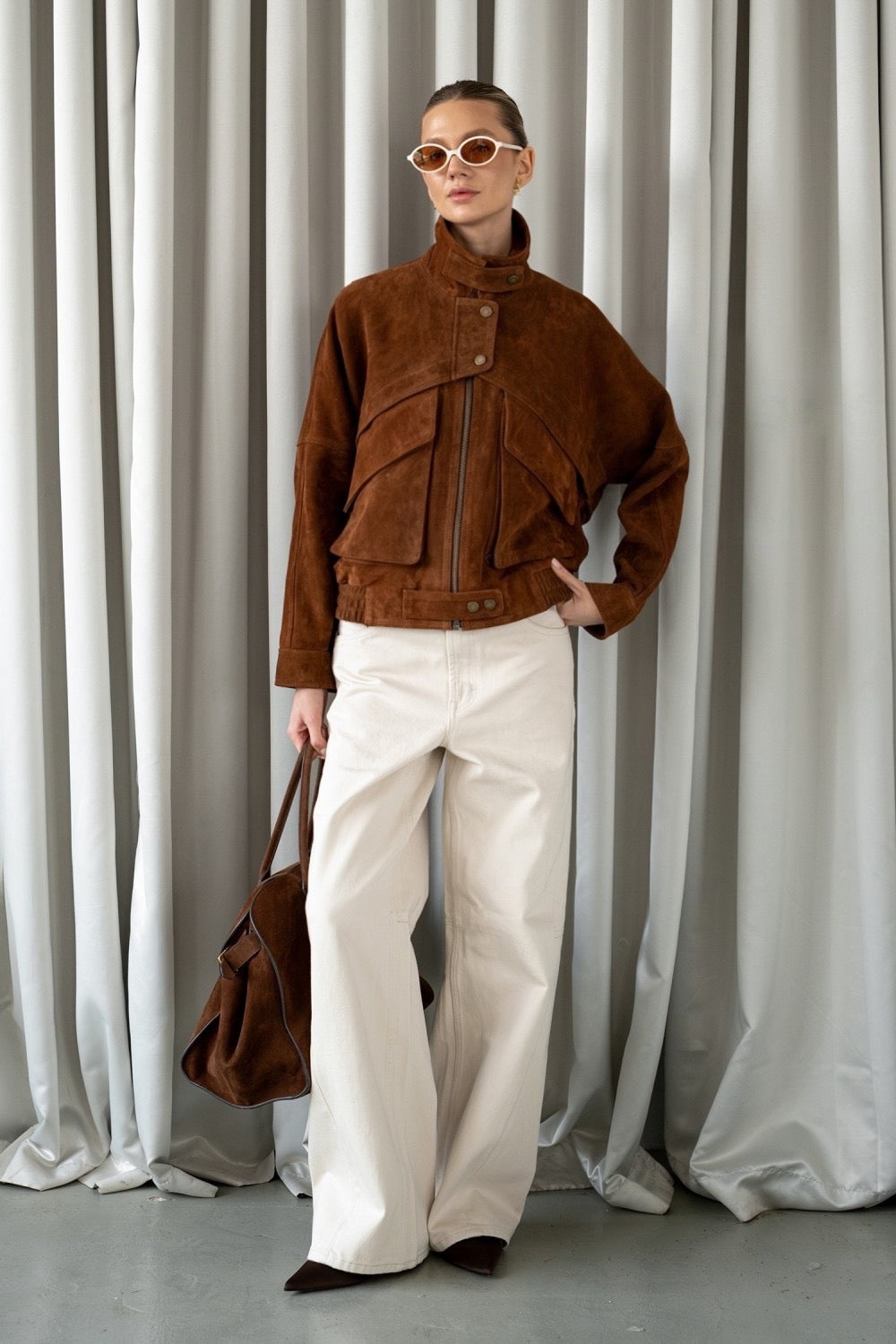 Elegant woman in cropped brown suede jacket, styled with wide-leg trousers and tortoiseshell sunglasses.