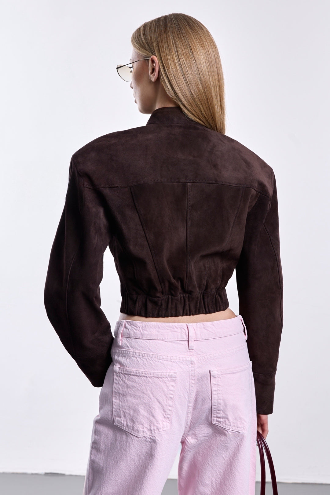 A woman showcases a luxurious cropped suede jacket in rich espresso, with strong shoulder tailoring, a high neckline, and a hidden closure for a refined, modern look.