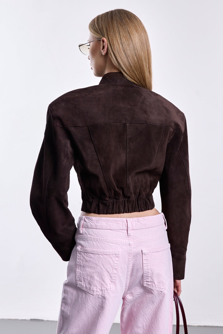 A woman showcases a luxurious cropped suede jacket in rich espresso, with strong shoulder tailoring, a high neckline, and a hidden closure for a refined, modern look.