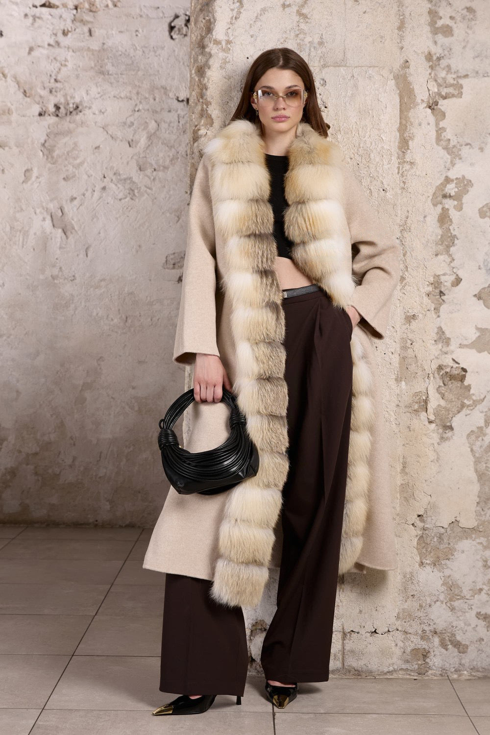 A woman wearing a beige cashmere coat with a gold fox fur trim, featuring a cinched waist and tailored silhouette for an elegant, feminine look.