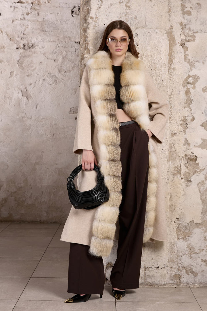 A woman wearing a beige cashmere coat with a gold fox fur trim, featuring a cinched waist and tailored silhouette for an elegant, feminine look.