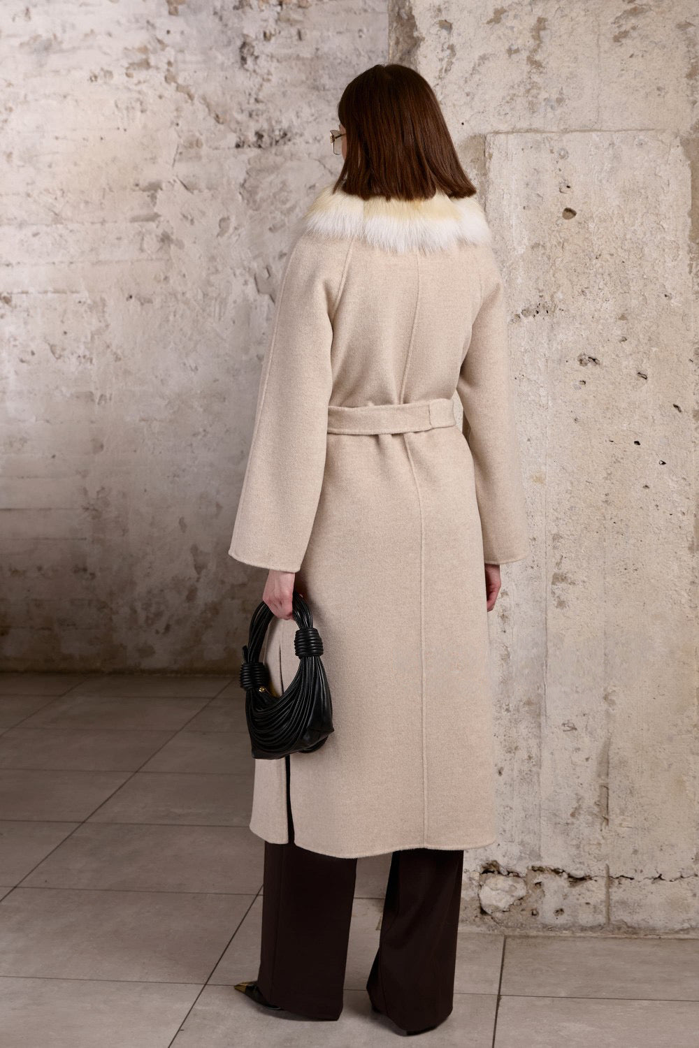 A woman wearing a tailored beige coat made from 100% cashmere, complete with a belted waist and rich gold fox fur trim for a timeless and elegant winter style.