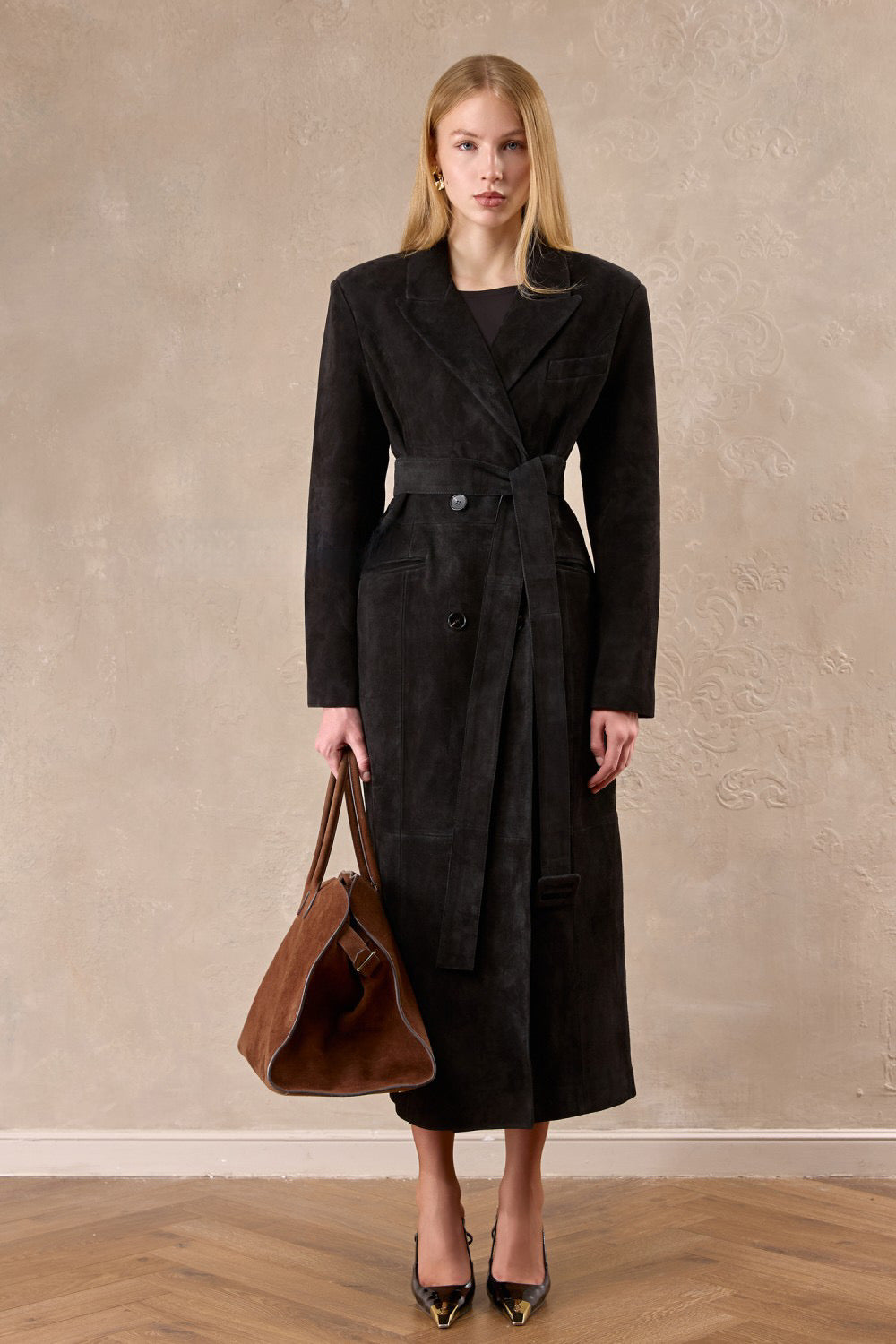 A woman wears a long dark brown suede coat with wide shoulders and a cinched belt, showcasing timeless elegance and strong tailoring.