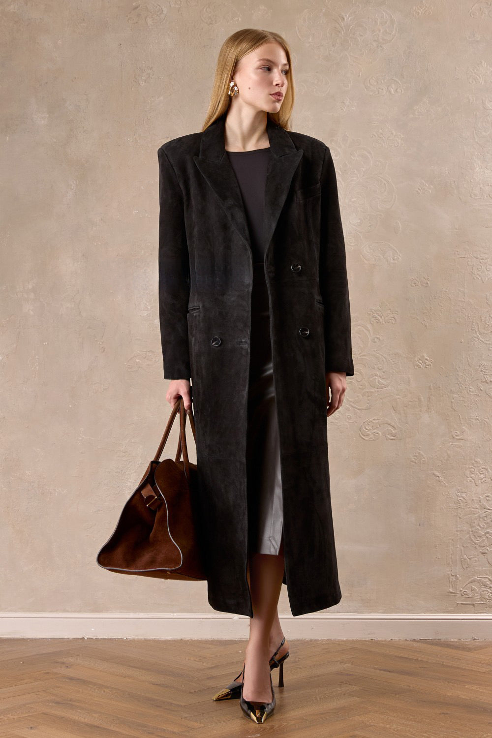 A woman models a premium dark brown suede coat, complete with sharp shoulders and a belted waist for a sophisticated, commanding look.