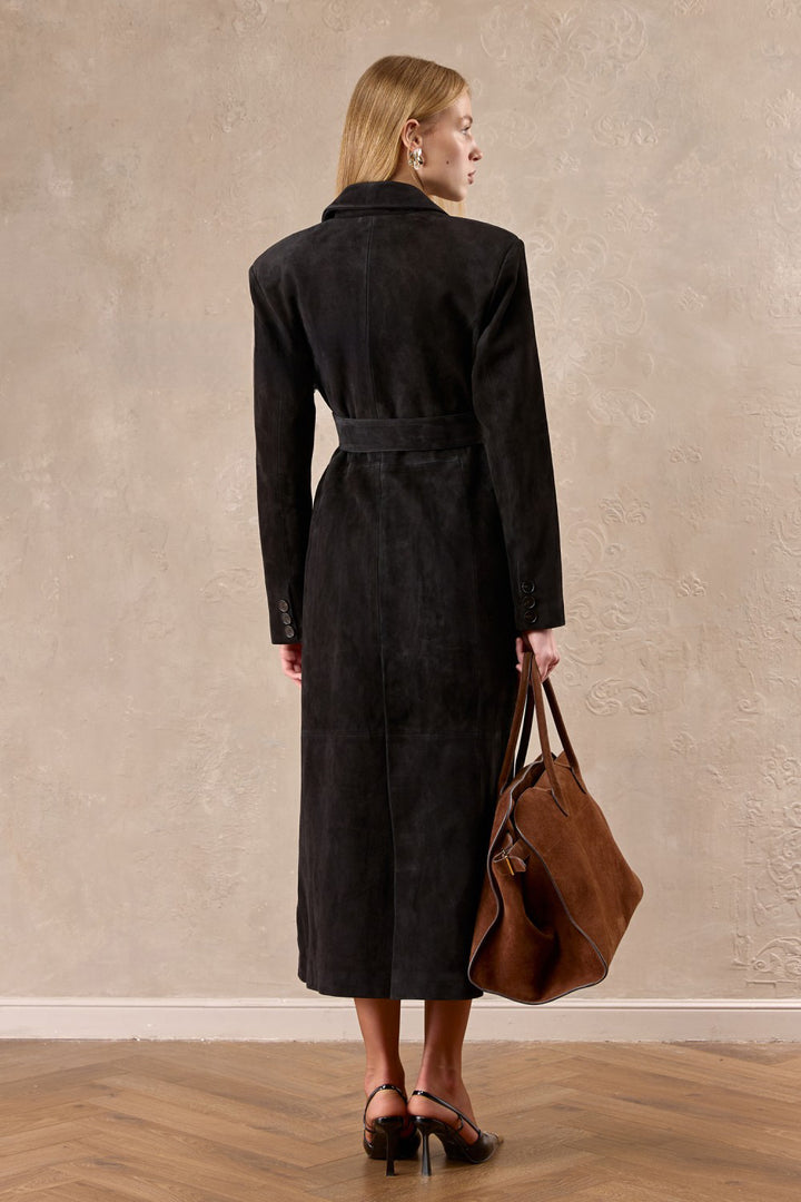 A woman in a long tailored suede coat radiates quiet confidence, the dark brown tone adding warmth and polish to her ensemble.