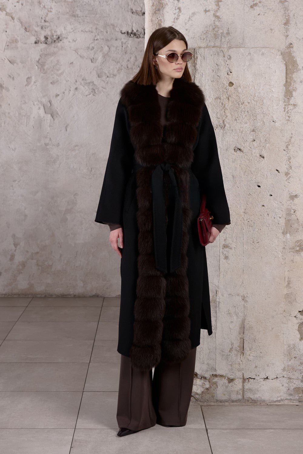 A woman wears a luxurious black cashmere coat, cinched at the waist with a belt and adorned with soft brown fox fur, creating a flawlessly elegant silhouette.