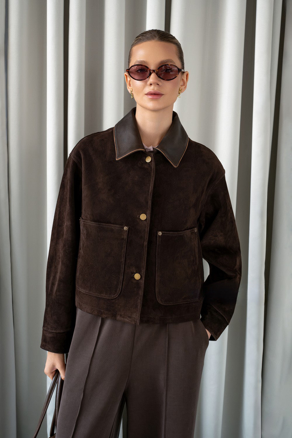 A woman wears an ultra-trendy brown suede leather jacket with a lapelled collar and front flap pockets, showcasing a structured and polished silhouette.