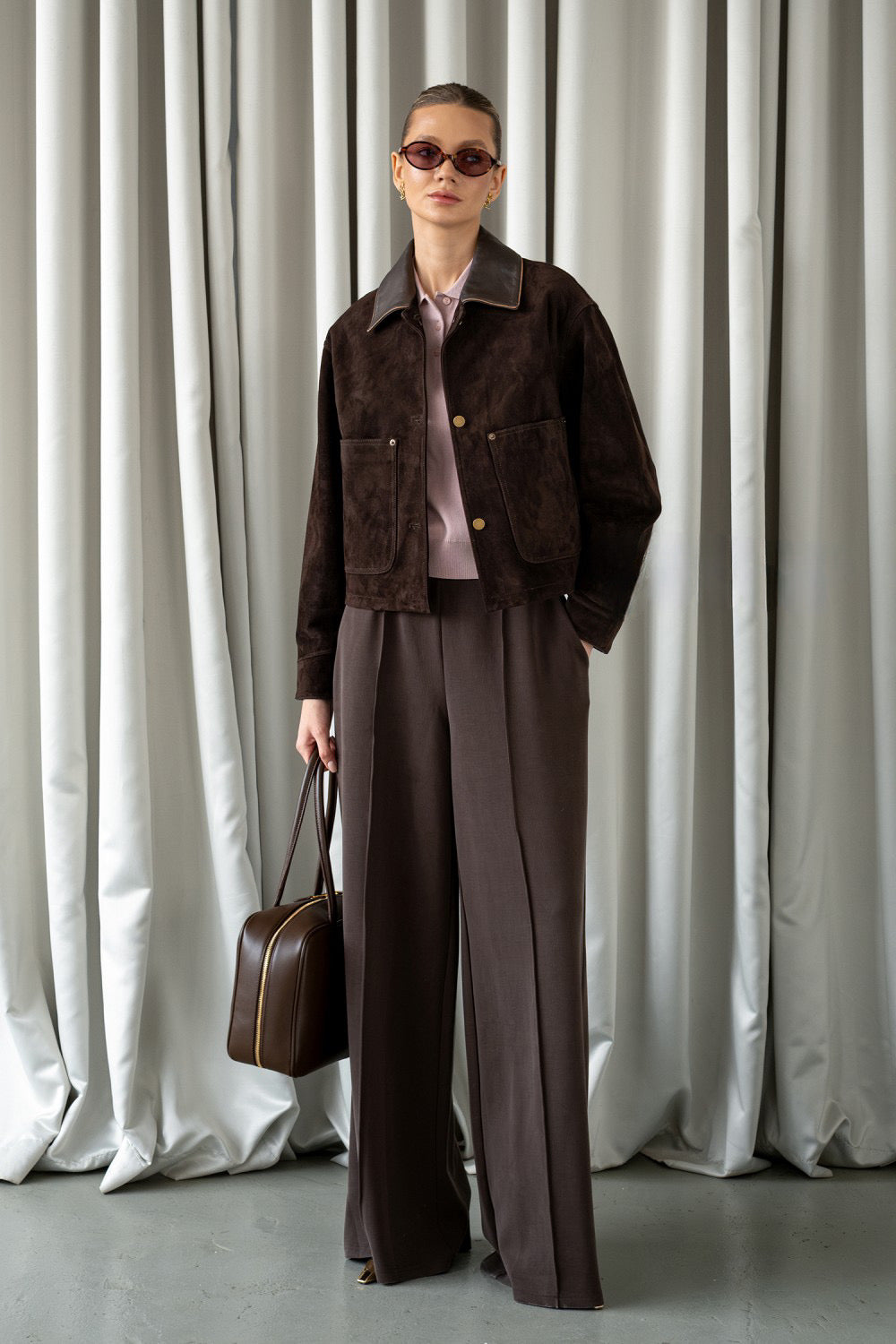 Stylishly dressed, a woman models a brown suede leather jacket featuring a soft texture, lapelled collar, and flattering structured fit.