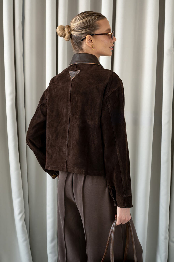 A woman is seen wearing a brown suede leather jacket with front flap pockets and a lapelled collar, combining depth, sophistication, and modern style.